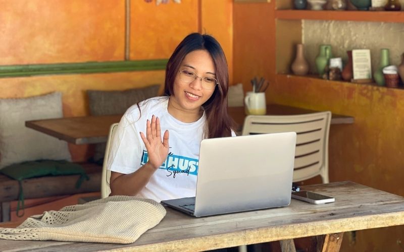 Female virtual assistant from The Side Hustle Squad waving during an online meeting at a cozy café workspace.