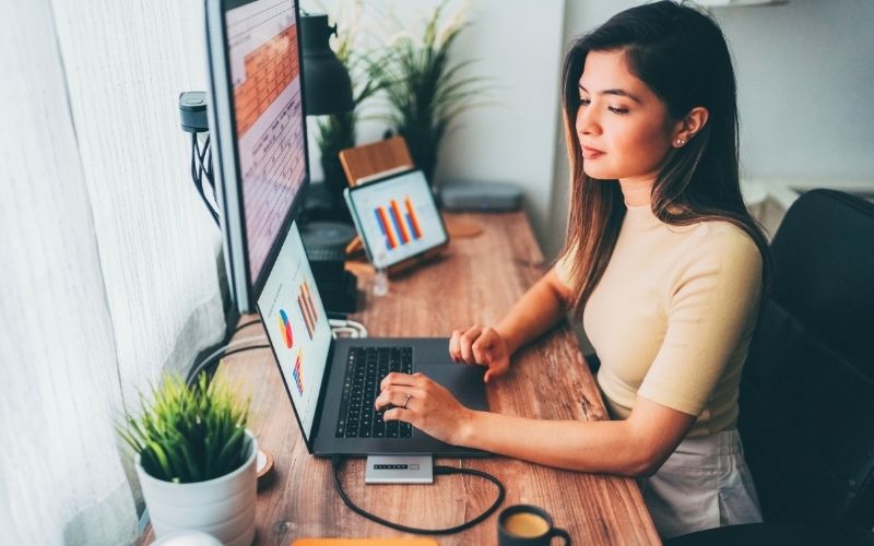 Focused virtual assistant analyzing charts and data on dual monitors while working from a home office.