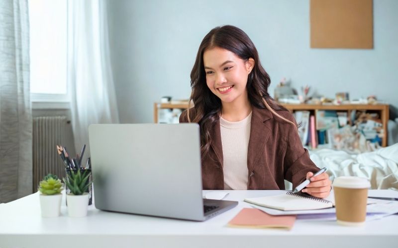 Smiling virtual assistant in a home office taking notes while attending an online meeting on a laptop.